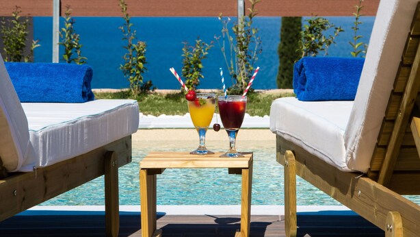 Zona de piscina con dos sillas de descanso, una mesa pequeña con dos bebidas y vista al mar.
