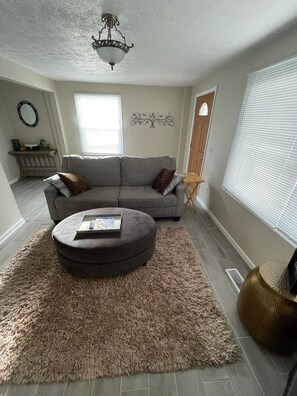 A living room with a grey sofa, a round ottoman, and a fluffy rug. There is a ceiling light fixture and a decorative wall piece. - Fun getaway house with arcade. Cedar Point/Kalahari/Sports Force Park/Summit Rac (Norwalk)