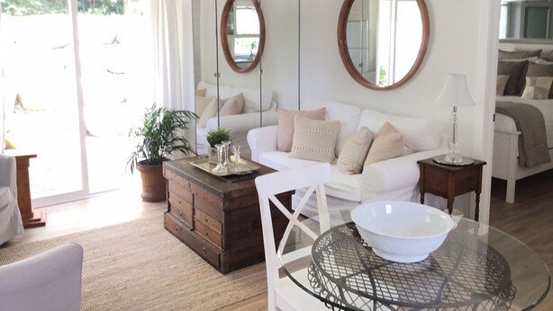 A modern living room with a white sofa, a wooden coffee table, and a glass-top table with a bowl. There are two round mirrors on the wall, a potted plant, and a sliding glass door leading outside. - One bedroom fully equipped Mahone Bay luxury cottage (Mahone Bay)