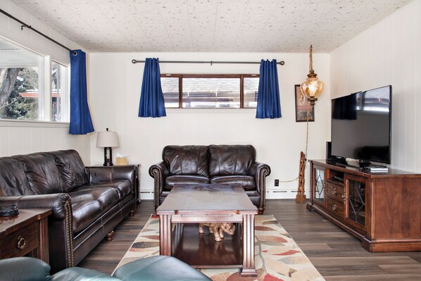 A living room with a brown leather sofa, a wooden coffee table, a TV on a wooden stand, and a window with blue curtains.