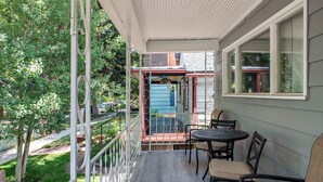 Eine überdachte Veranda mit Tisch und Stühlen, einem Geländer und Blick auf ein blaues Haus im Hintergrund.
