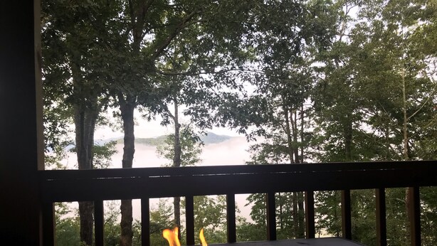 Ein Lagerfeuer mit Flammen, umgeben von einer Holzdielen-Terrasse mit Blick auf eine bewaldete Landschaft, einen See und entfernte Berge.