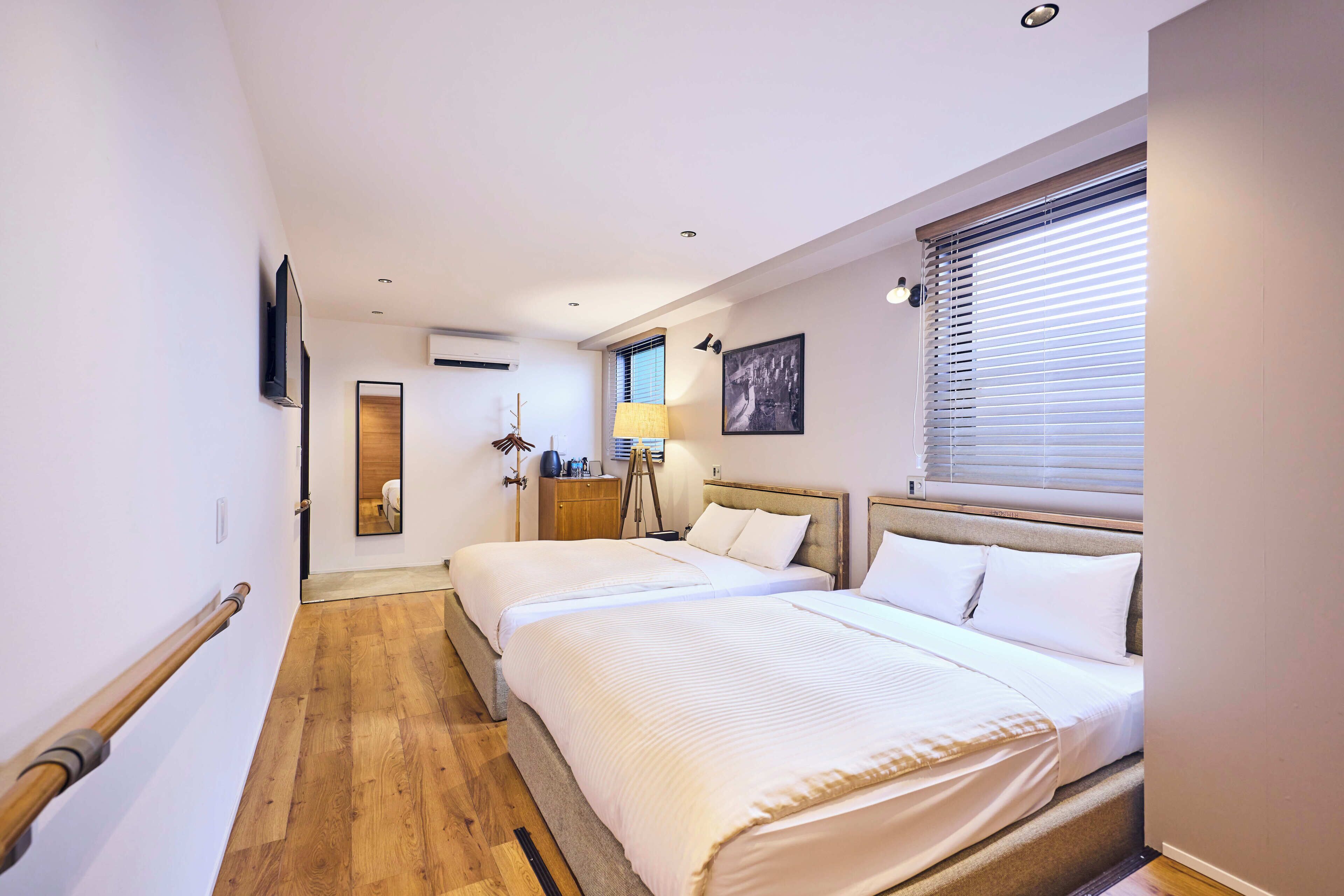 A hotel room with two beds, a wooden floor, a flat-screen TV mounted on the wall, a window with blinds, and a bedside table with a lamp.