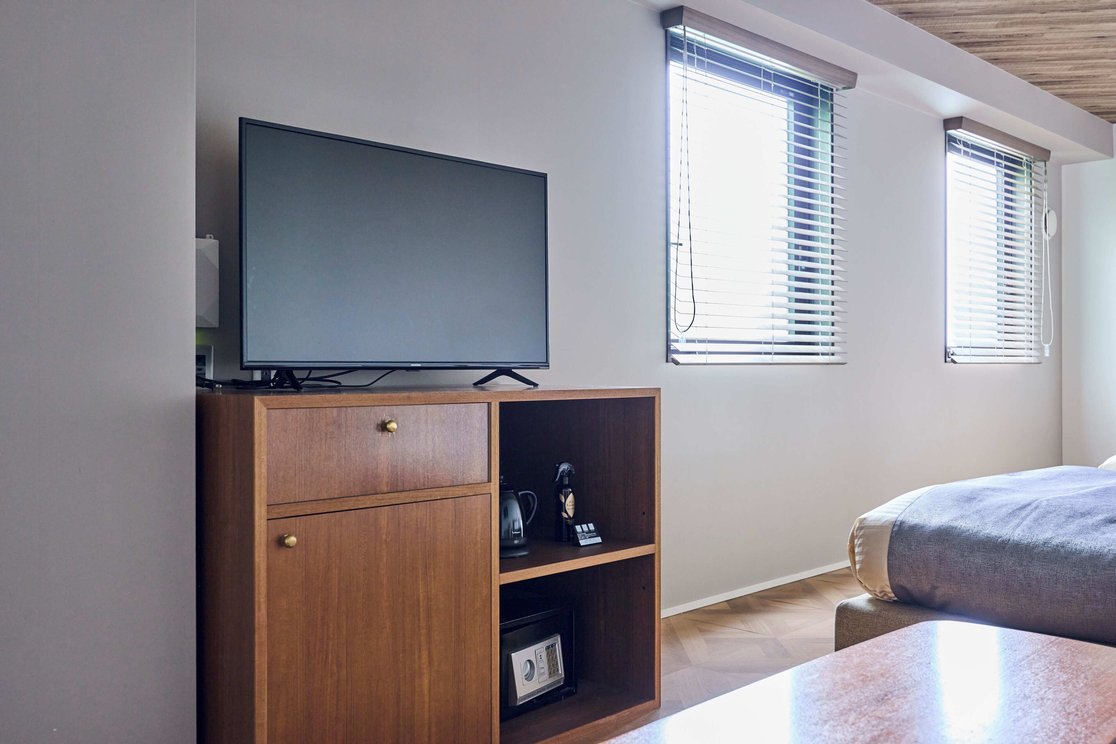 A bedroom with a TV on a wooden cabinet, a bed, and two windows with blinds.
