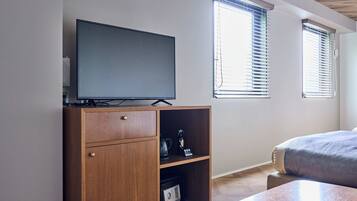 A bedroom with a TV on a wooden cabinet, a bed, and two windows with blinds.