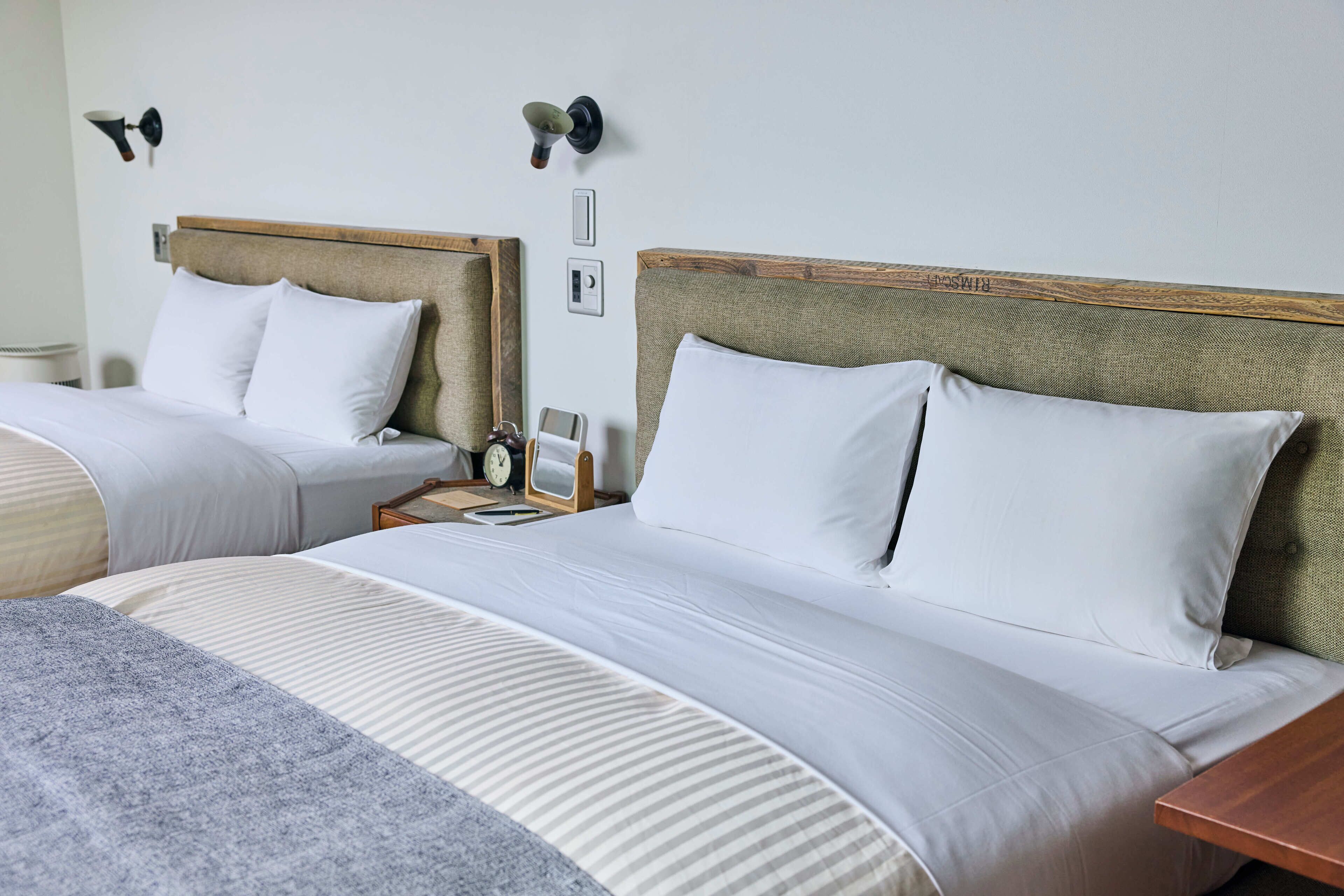A hotel room with two beds, each with a wooden headboard, white bedding, and a bedside table with a clock and a small table lamp.