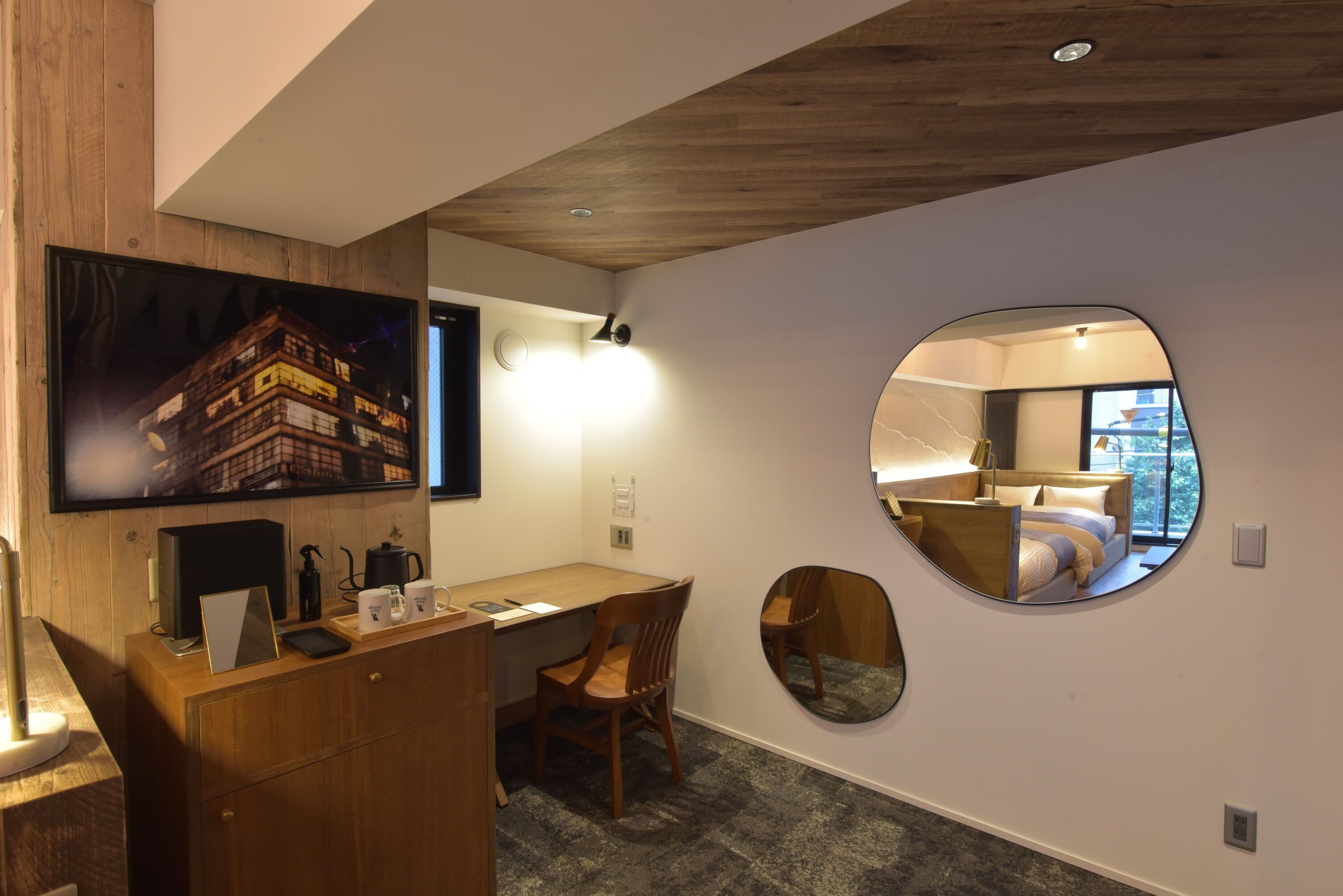 Habitación de hotel moderna con un escritorio de madera, una silla, un televisor de pantalla plana y un espejo redondo que refleja la cama y la ventana.