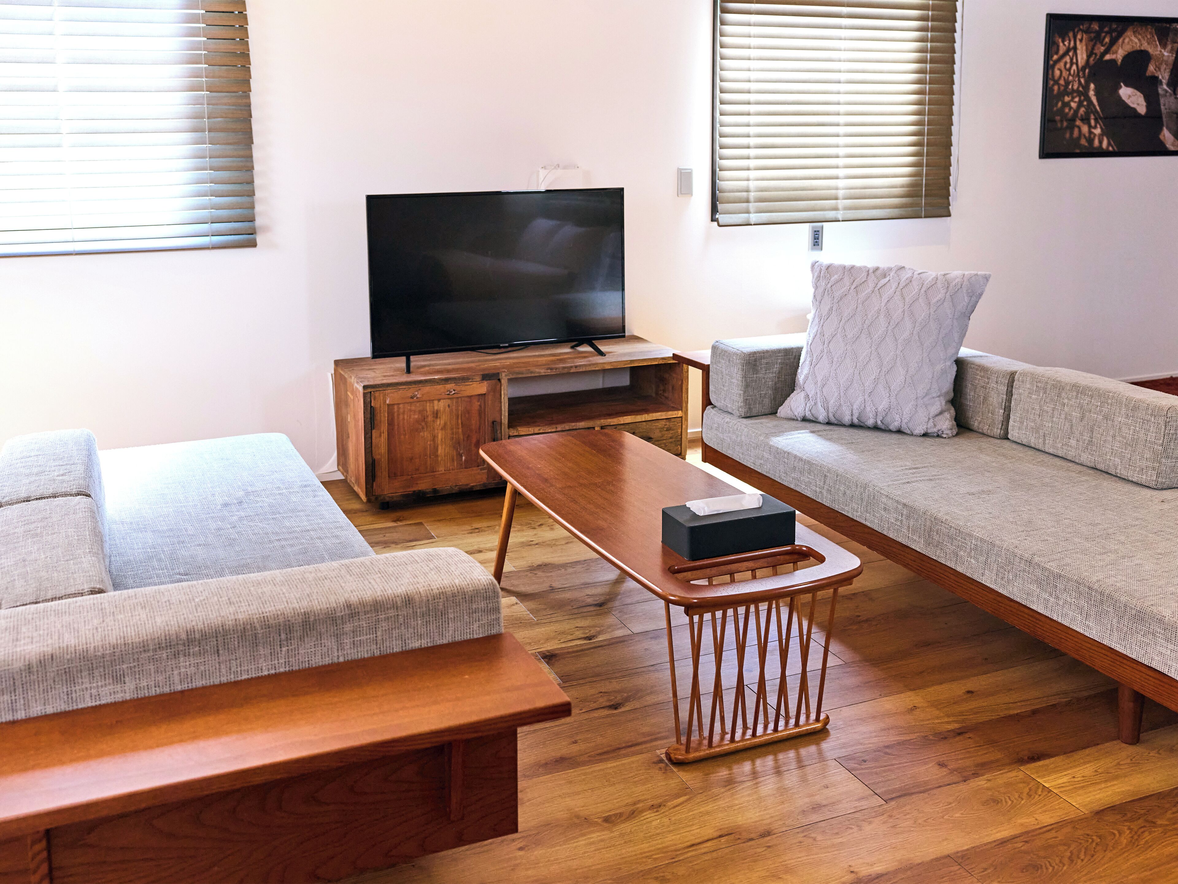 A living room with a wooden coffee table, a sofa, a television on a wooden stand, and a window with blinds.