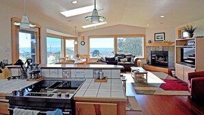 A modern kitchen with a central island, a dining area with a red rug, and a living room with a fireplace and large windows. - The Waves House! Breathtaking Ocean View, Hot Tub, King Beds, Dog-Friendly. (Waldport)