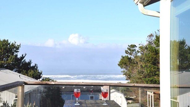 A wooden table with two glasses of red wine, overlooking a coastal view with the ocean and trees in the background. - The Waves House! Breathtaking Ocean View, Hot Tub, King Beds, Dog-Friendly. (Waldport)