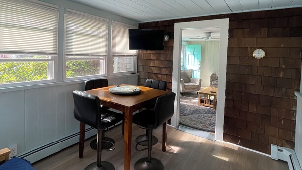 A room with a wooden dining table, four chairs, a flat-screen TV mounted on the wall, and a view through an open door to a dining area. - Cape Cod Beach Cottage, only 5 minute walk to Glendon beach & local restaurants (Dennis)