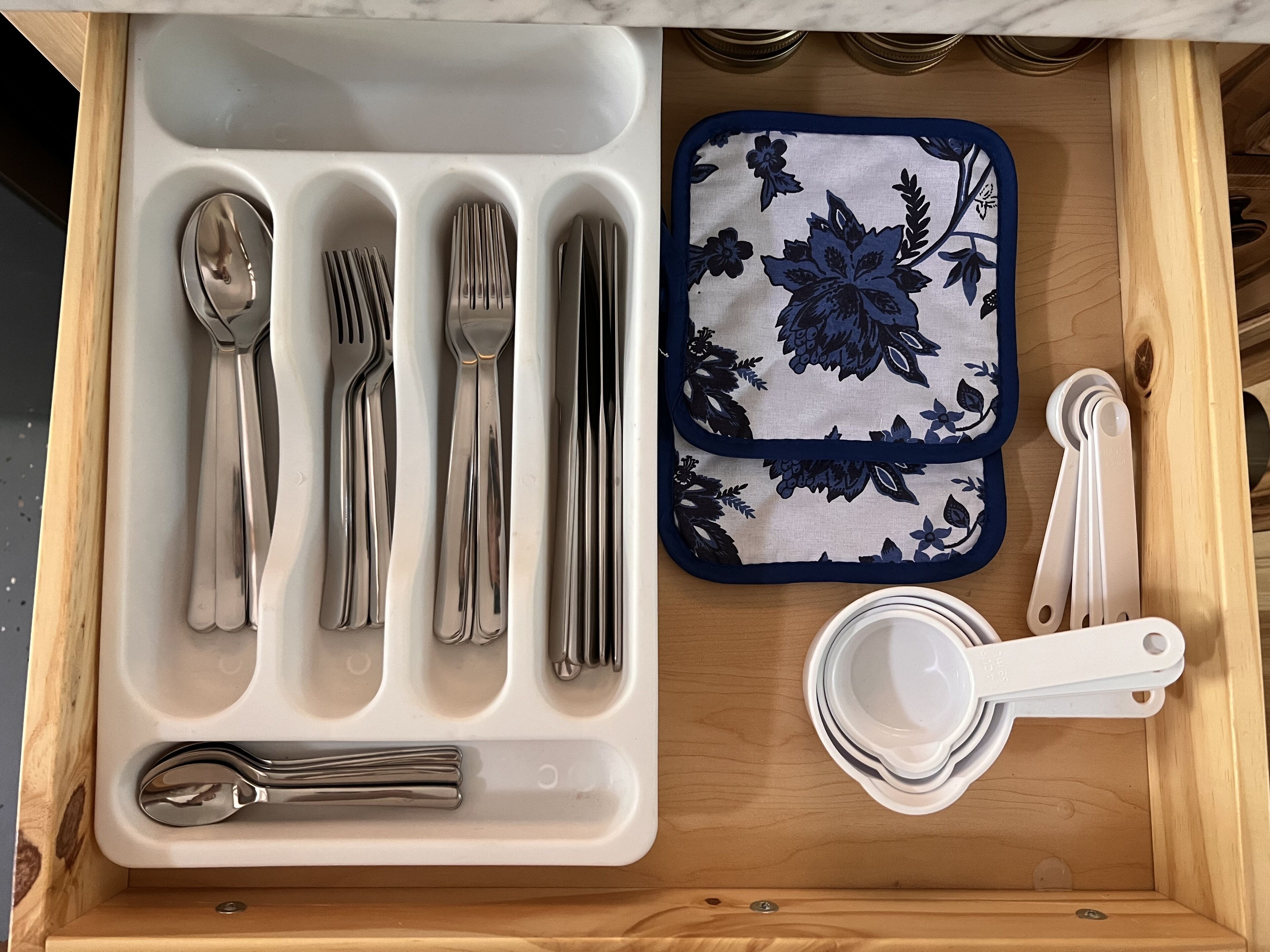A drawer with neatly organized silverware, a floral-patterned napkin holder, and measuring spoons.