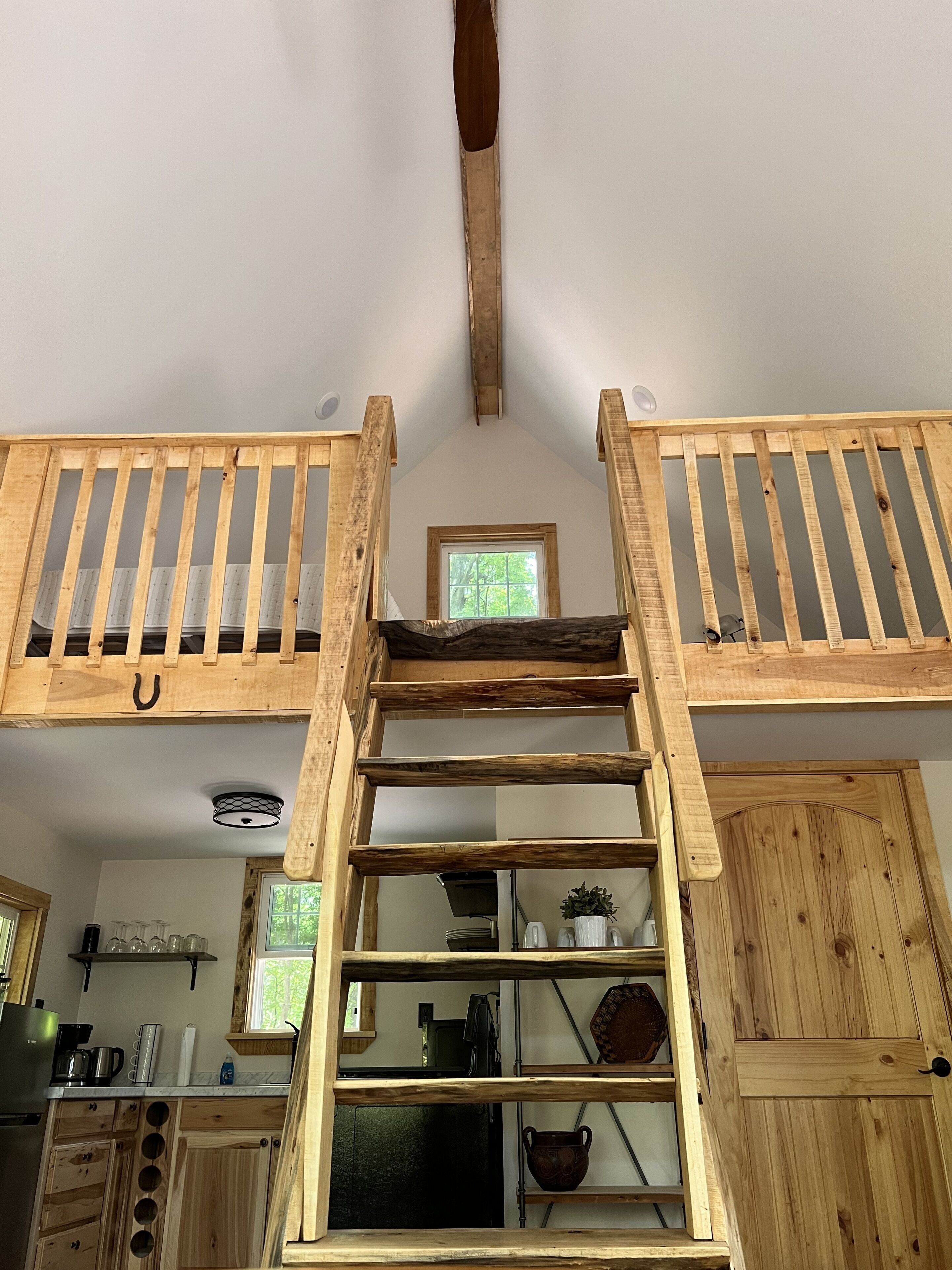Dormitorio estilo loft con una escalera de madera que lleva a una zona de descanso superior, una ventana pequeña y una cocina a la vista abajo.
