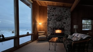 A cozy room with a stone fireplace, wooden walls, a large window overlooking a snowy landscape, and a comfortable seating area.