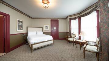A hotel room with a large bed, a desk with a lamp, two chairs, a small table, and a door.