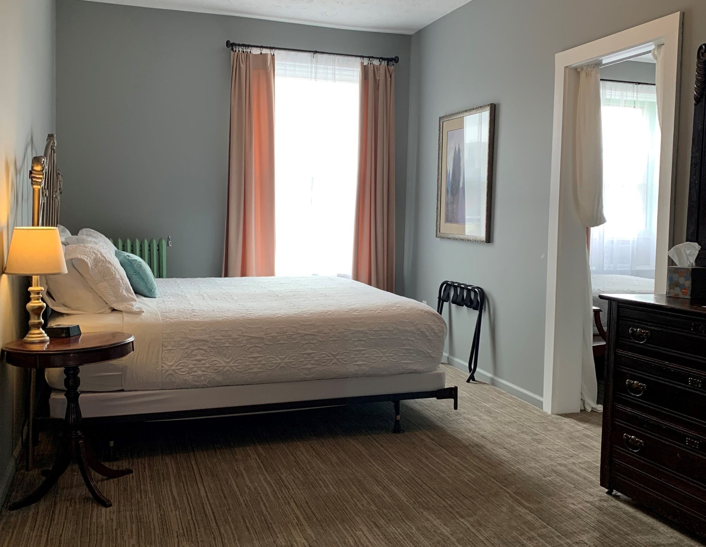 A neatly arranged bedroom with a bed, bedside table, lamp, radiator, and a dresser.