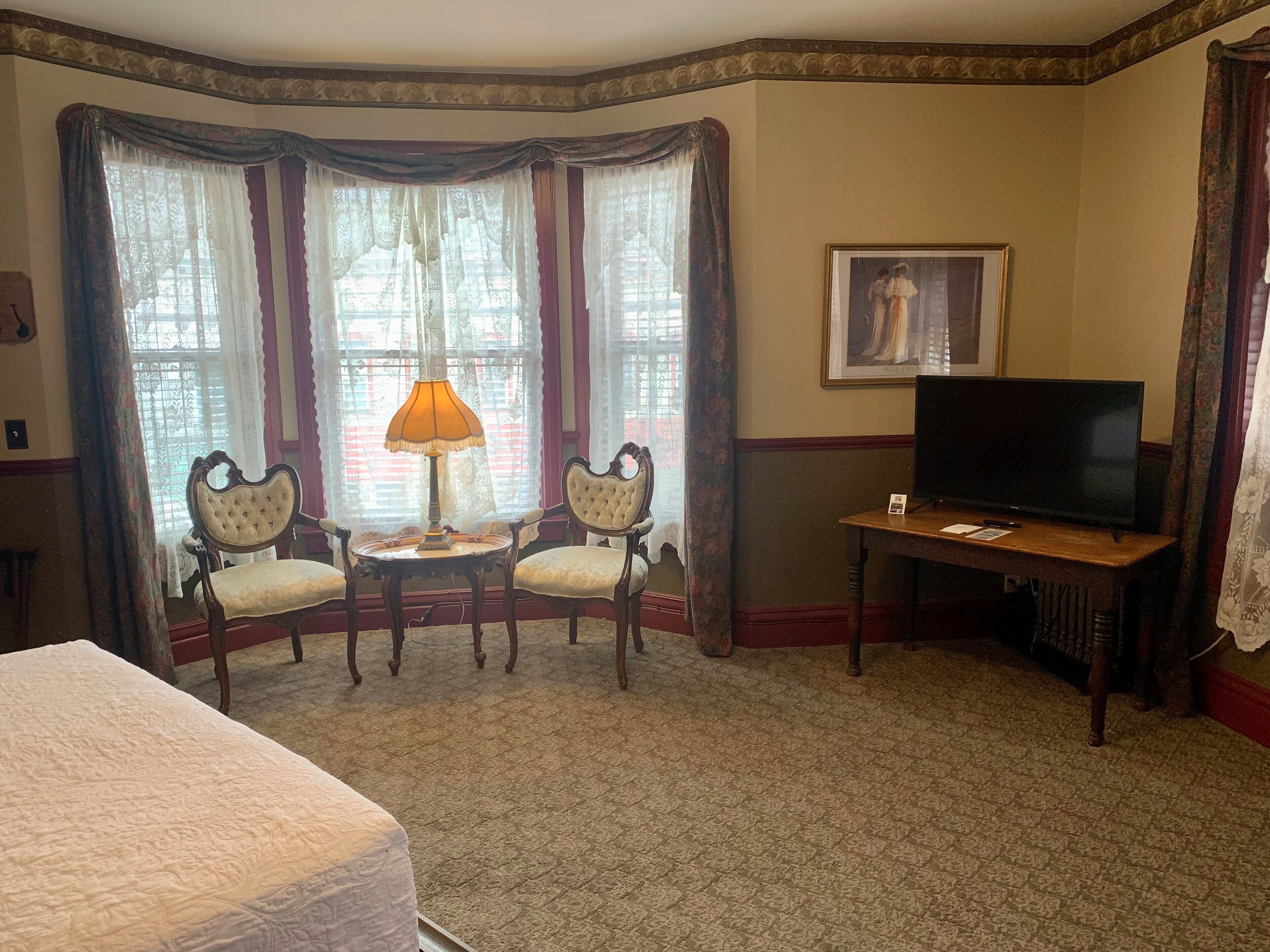 Habitación de hotel con una cama grande, dos sillones, una mesita con una lámpara, un televisor y un cuadro en la pared.