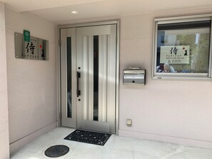 A hotel entrance with a metallic door, a manhole cover, a window with a view, and a wall-mounted sign. - Samurai Hills (Kyoto)