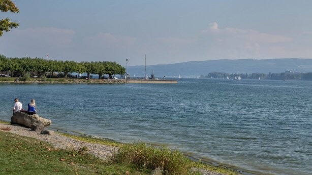 Vista de un lago con muelle, agua tranquila y colinas al fondo.
