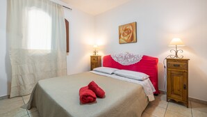 Un lit bien fait, recouvert d’une couvre-lit beige et agrémenté de deux serviettes rouges, deux lampes de chevet, une table de chevet en bois, une tête de lit rouge et un tableau encadré au mur.