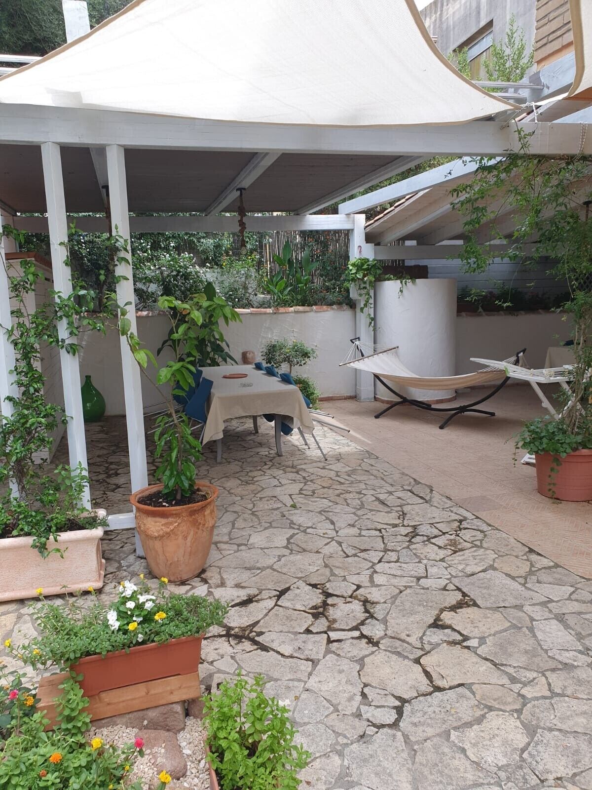 Un porche cubierto con una mesa, sillas, plantas en macetas y una hamaca.