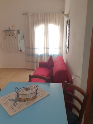 Una habitación con una mesa azul, un sofá rojo, un camino de mesa estampado y una ventana con cortinas.