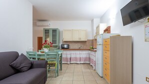 Un ambiente compatto con divano, tavolo da pranzo, sedie, angolo cottura dotato di mobili e forno a microonde, e TV a parete.