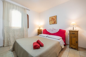 Un lit bien fait, recouvert d’une couvre-lit beige, avec deux oreillers et deux serviettes rouges. On y trouve deux tables de chevet avec des lampes, une petite table en bois et un tableau représentant une rose, accroché au mur.