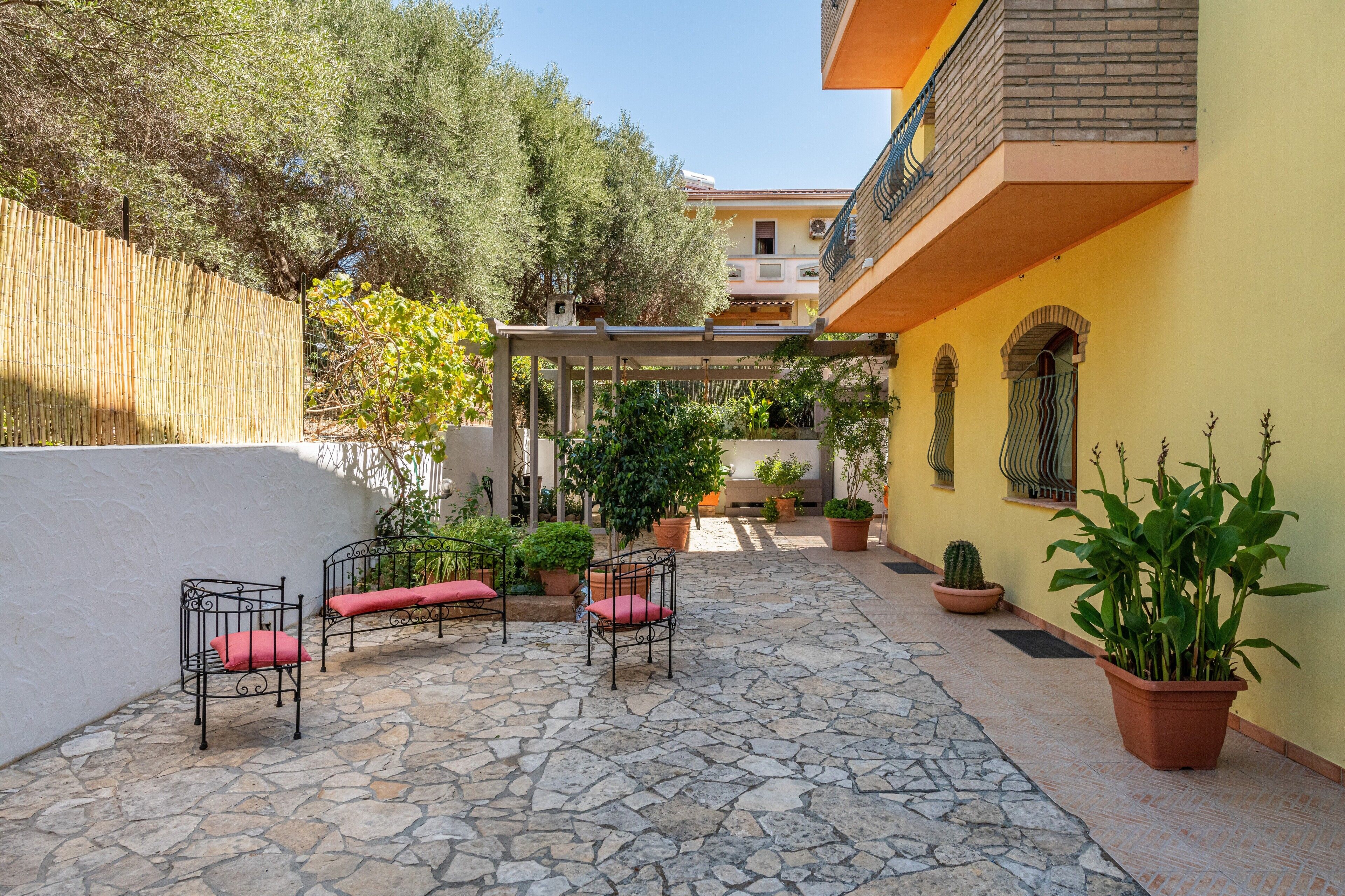 Patio esterno con pavimento in pietra, sedie in metallo con cuscini rossi, piante in vaso e un edificio giallo con un balcone.