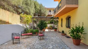 Patio esterno con pavimento in pietra, sedie in metallo con cuscini rossi, piante in vaso e un edificio giallo con un balcone.
