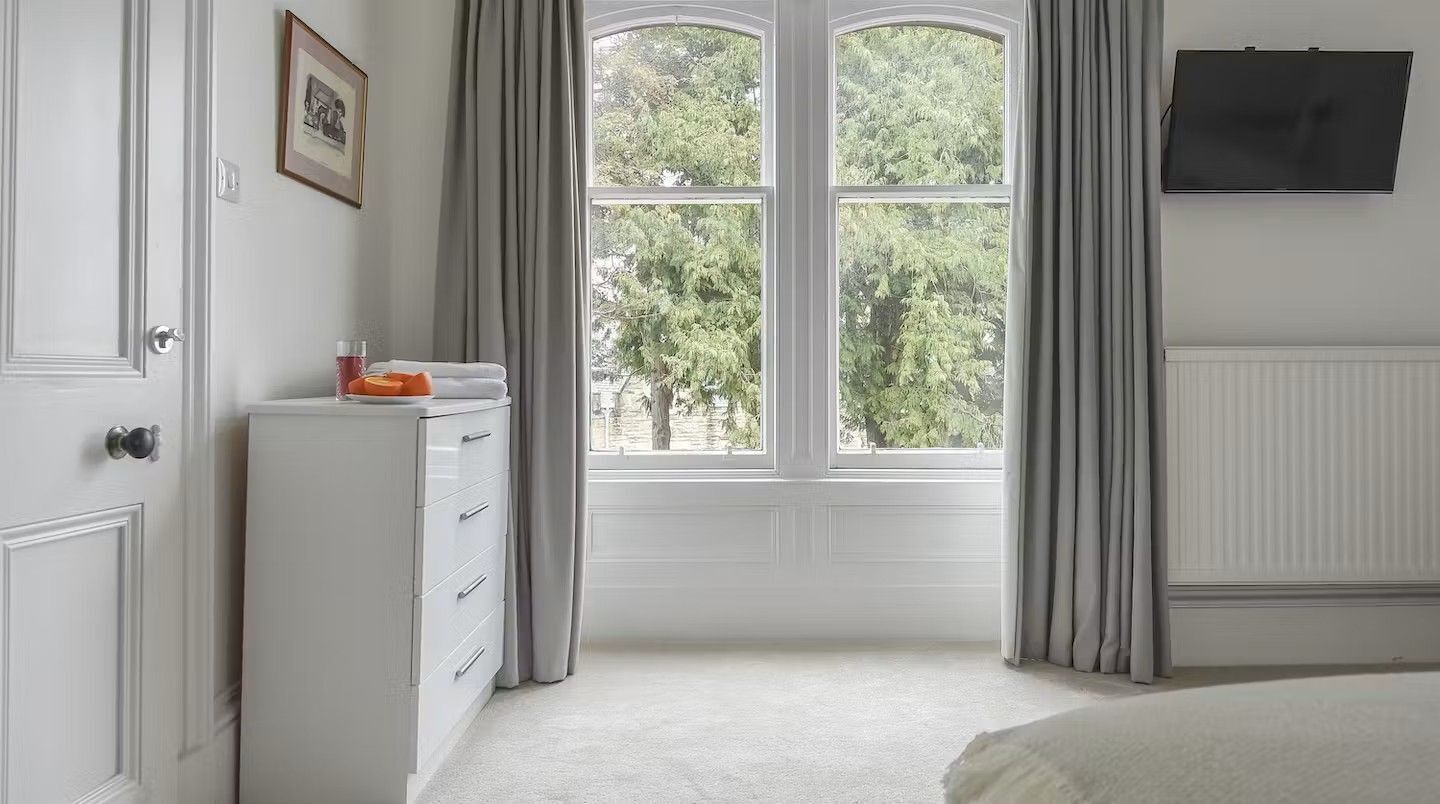 Een slaapkamer met een groot raam, een bed, een dressoir, een televisie en een ingelijst schilderij aan de muur.