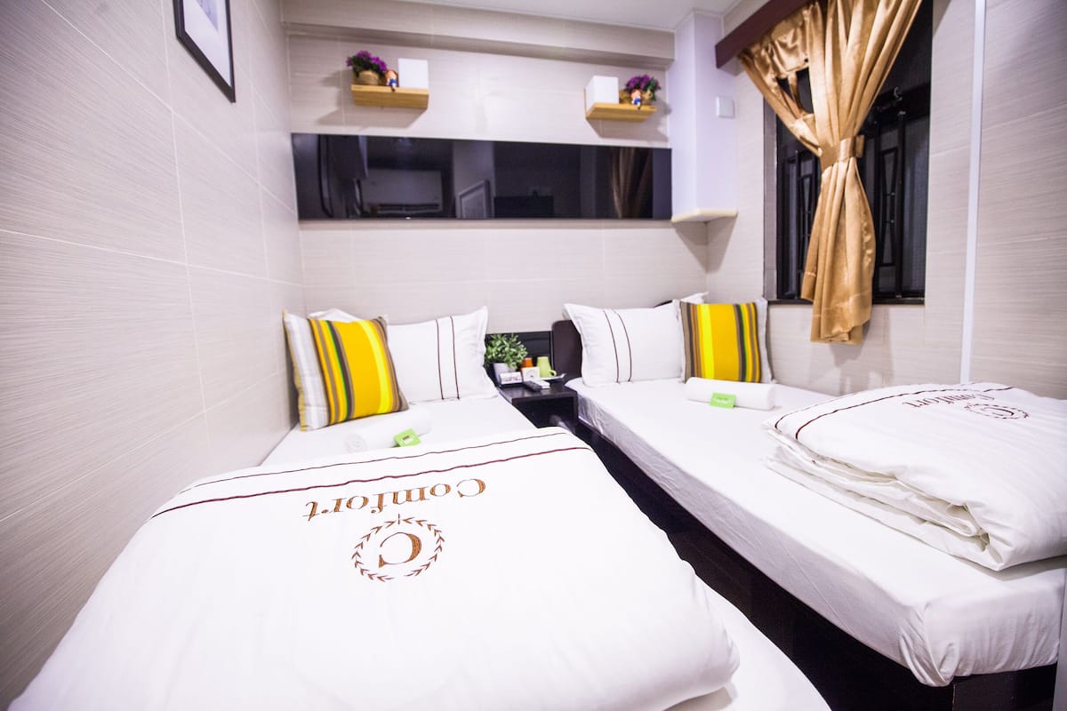 Quarto de hotel com duas camas, cada uma com lençóis brancos e um logotipo no edredom. Há uma pequena mesa com uma planta e um abajur entre as camas.