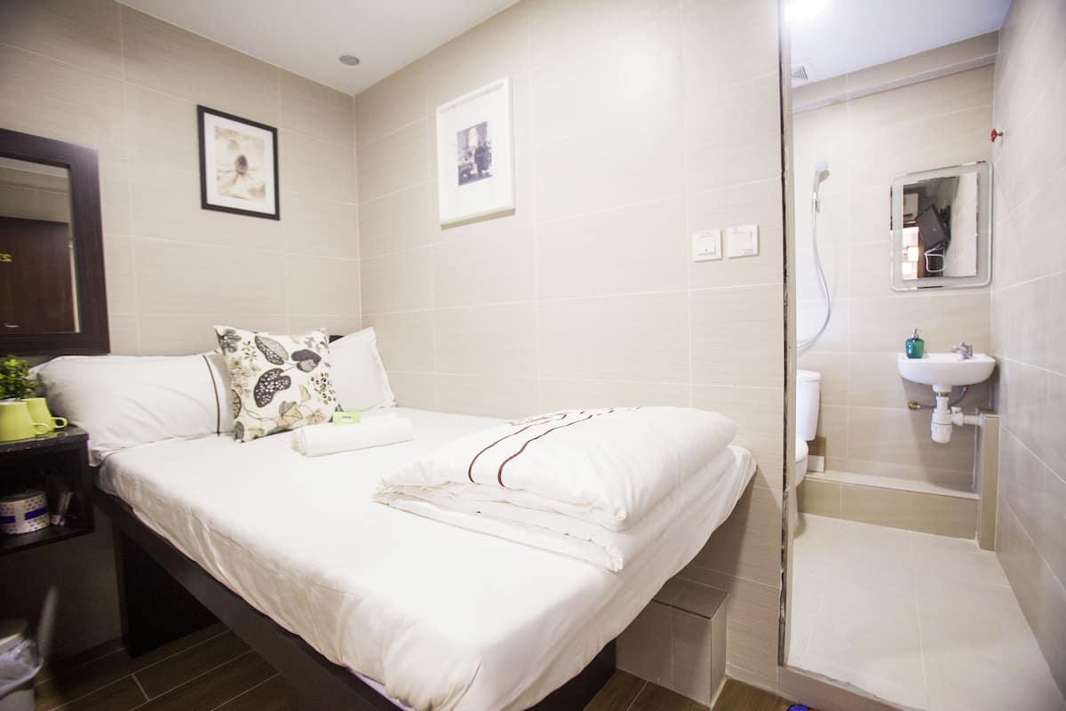 Quarto de hotel com uma cama, uma mesinha de cabeceira com uma planta e um banheiro com chuveiro e pia.