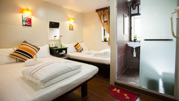 Una habitación de hotel con dos camas, una mesita de noche, un espejo, un televisor montado en la pared y un baño con lavamanos y ducha.