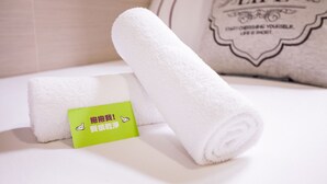 Una toalla blanca doblada con una etiqueta verde sobre una superficie blanca, junto a una toalla enrollada y un almohadón con la palabra 'LIFE'.