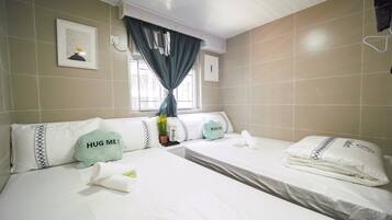 A hotel room with two beds, each with white bedding and decorative pillows, including one with "HUG ME!" text. There is a window with curtains, a wall-mounted light, and a framed picture above the window.