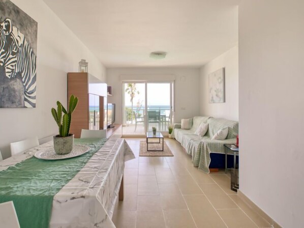 A spacious living area with a large window overlooking the sea, a dining table with a green tablecloth, and a sofa with a light-colored cover.