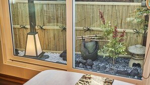 Quarto com uma grande janela com vista para uma cerca de bambu e um pequeno jardim com um candeeiro, uma bacia de pedra e uma casa de pássaros decorativa.