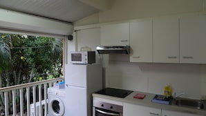 Een kleine keuken met een magnetron, oven en spoelbak. Er is een wasmachine en een balkon met uitzicht op groen.