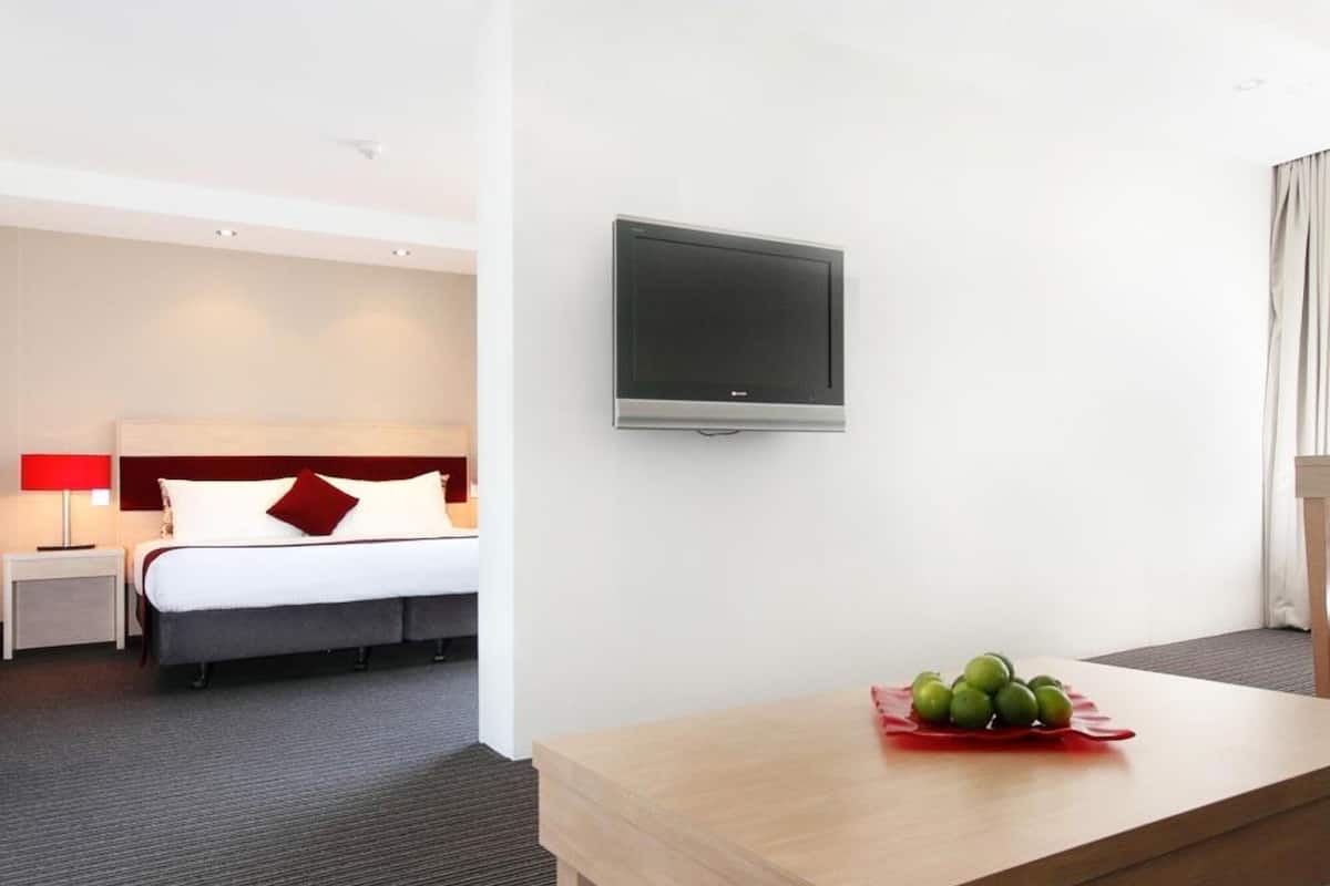 Habitación de hotel con cama, televisor montado en la pared, una mesa de madera con frutero y suelo alfombrado.