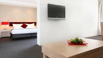 A hotel room with a bed, a TV mounted on the wall, a wooden table with a fruit bowl, and a carpeted floor.