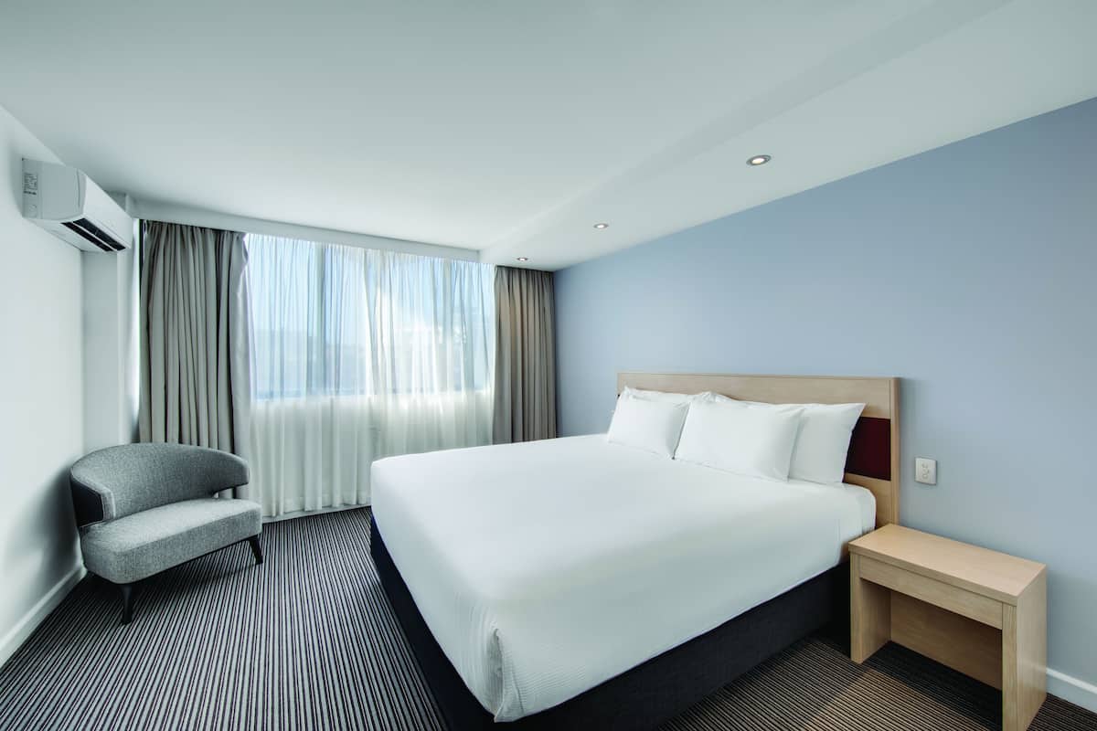 Habitación de hotel con una cama grande, una silla, una mesita de noche y una ventana con cortinas.