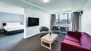 A hotel room with a bed, a TV, a sofa, a coffee table, and a view of the city. - Central Studio Hotel Sydney (Haymarket)