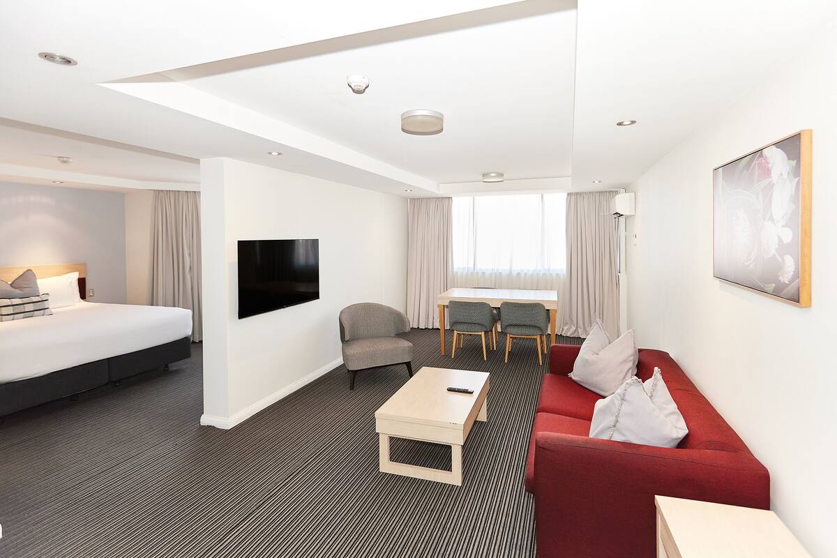 Una habitación de hotel con una cama, un sofá rojo, una mesa de centro, un televisor, un escritorio con sillas y un ventanal con cortinas.
