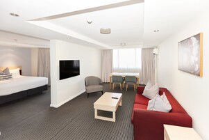 A hotel room with a bed, a red sofa, a coffee table, a TV, a desk with chairs, and a large window with curtains.