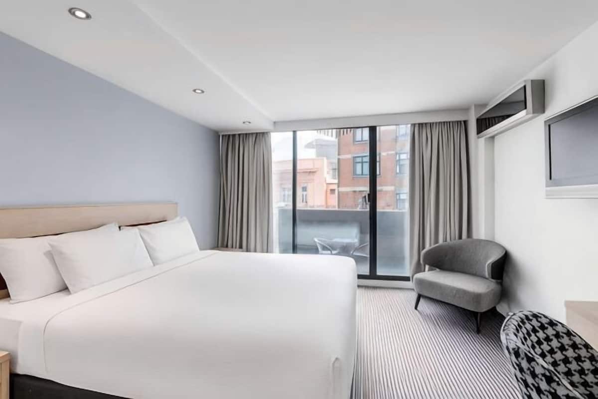 Habitación de hotel con una cama grande, una silla, un televisor y vistas a edificios por la ventana.