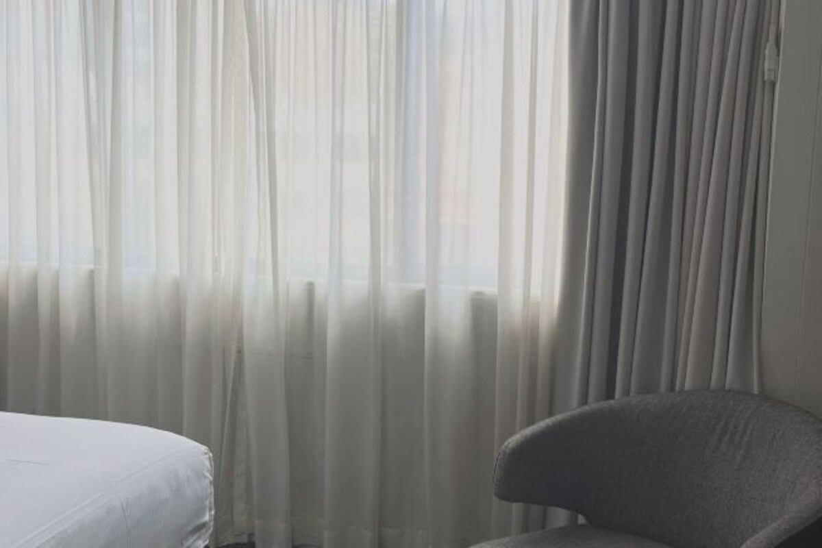 A room with a bed and a chair, curtains partially drawn, and a window with a view.