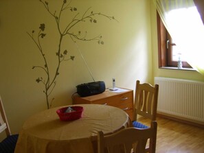 Een kamer met een ronde tafel, een rode mand, een radio, een raam met gordijnen en een muur met bloemendecoratie.
