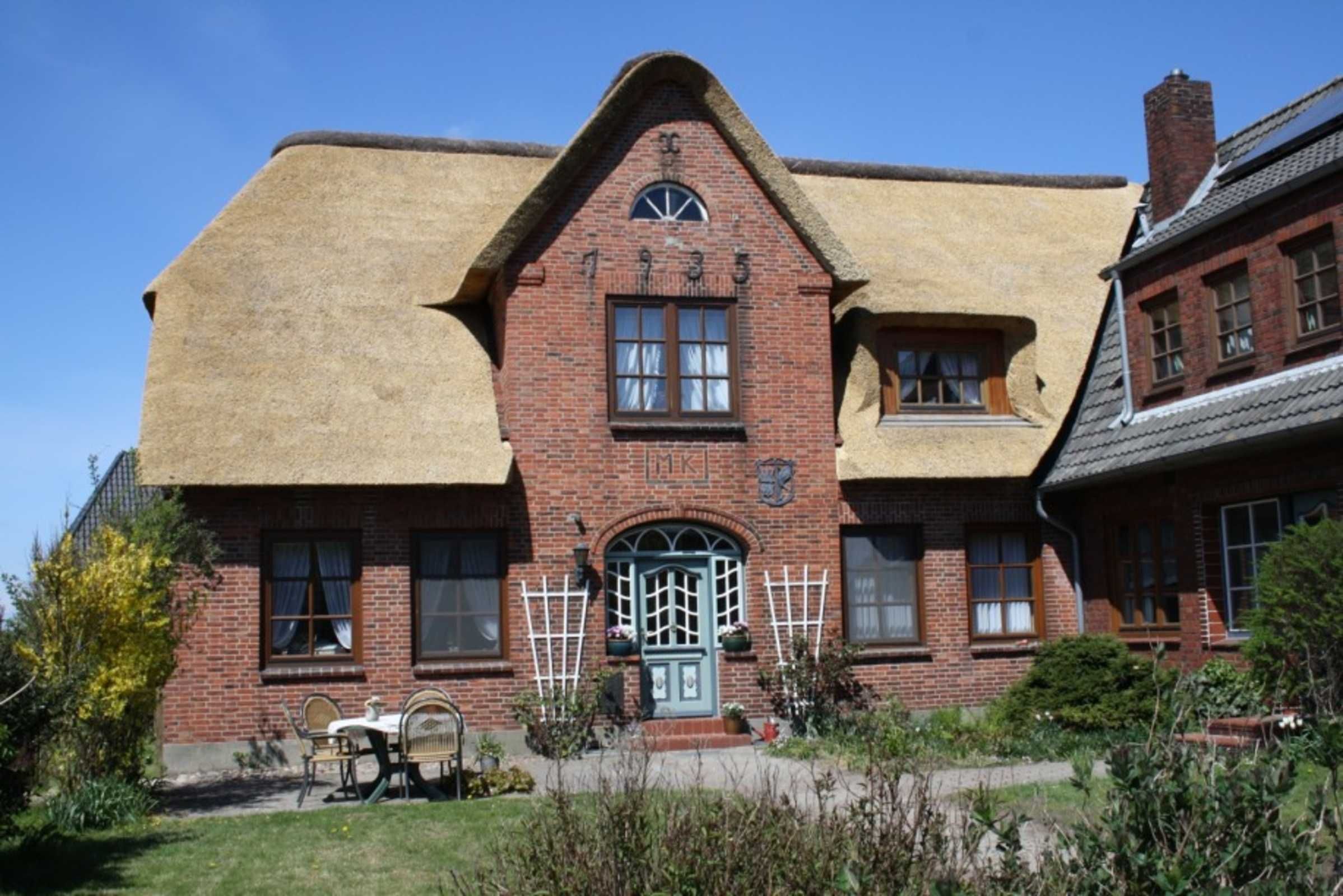A brick building with a green door and windows, a thatched roof, and a small garden with a table and chairs.
