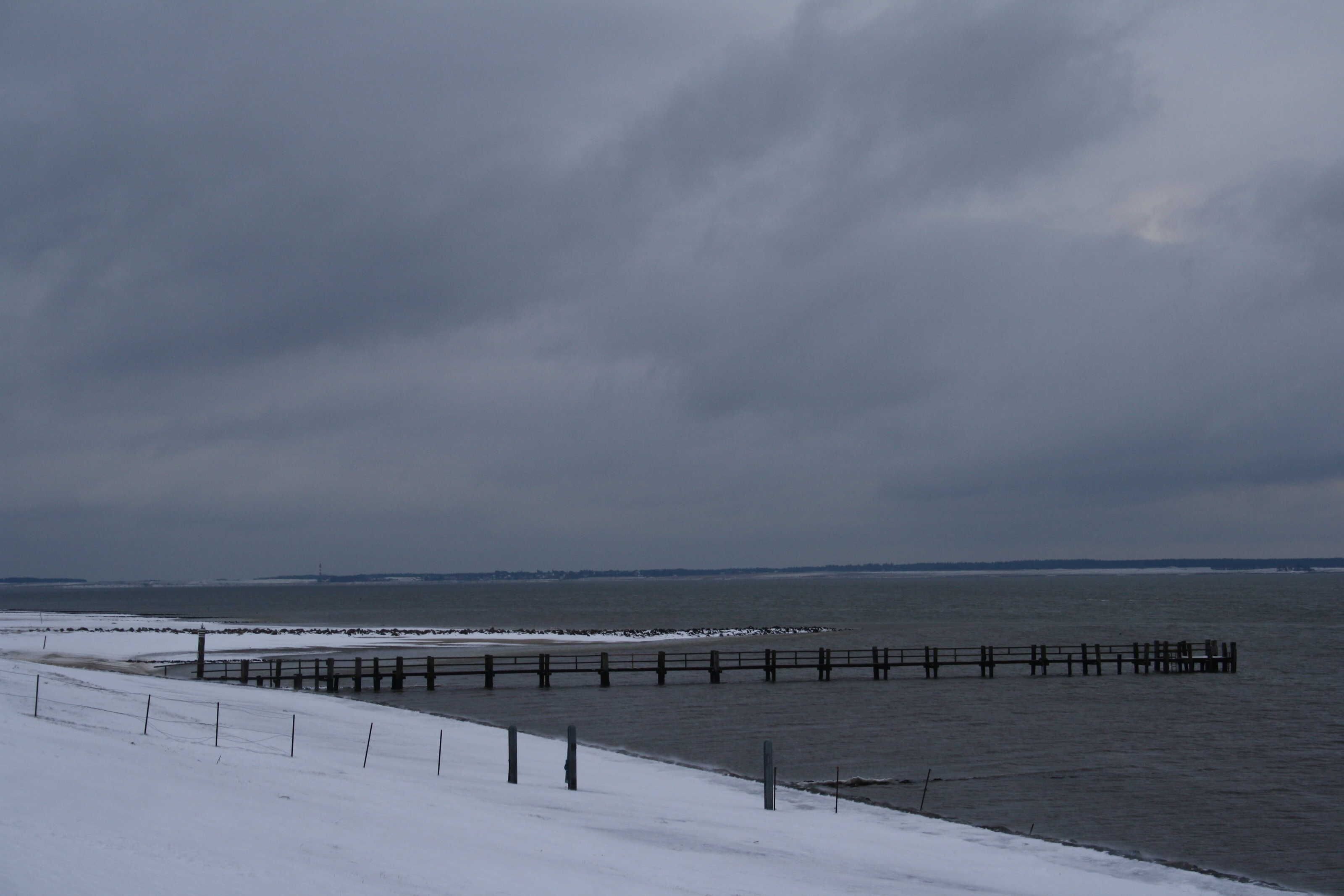 Eine Küstenansicht mit einem hölzernen Steg, der sich ins Wasser erstreckt, einem verschneiten Strand und einem bewölkten Himmel.